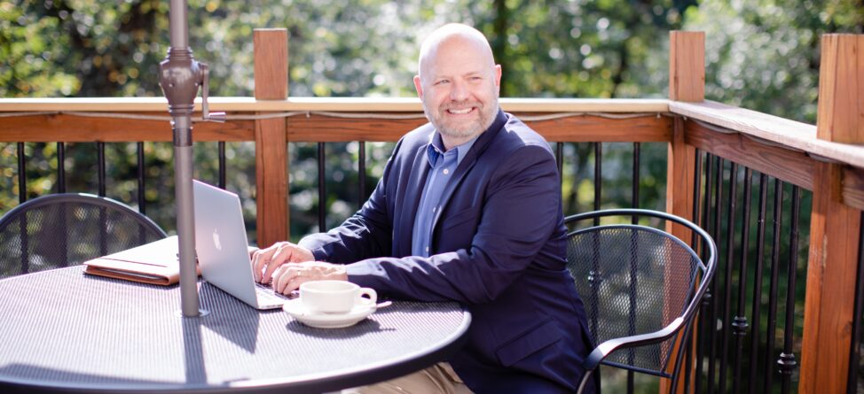 Financial planner Jim White smiling while working on a laptop outdoors at a sunny patio table